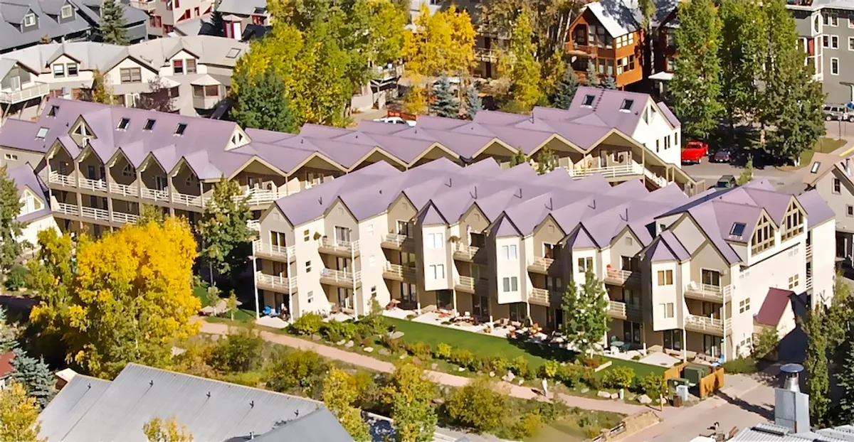 A vibrant row of buildings is set among lush greenery with a well-tended walking path in front. People can be seen strolling and relaxing outdoors, suggesting a lively but laid-back community atmosphere. The balconies invite morning coffee moments while the open space hosts small community gatherings.