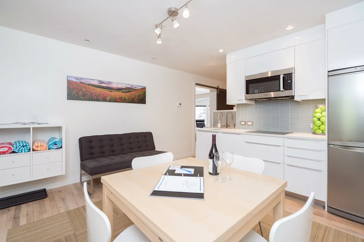 A welcoming interior scene with a dining table set with wine and glasses, modern kitchen appliances in the background, colorful artwork on the wall, and a comfortable sofa seating area. The layout feels tailored for casual gatherings and relaxed entertaining.