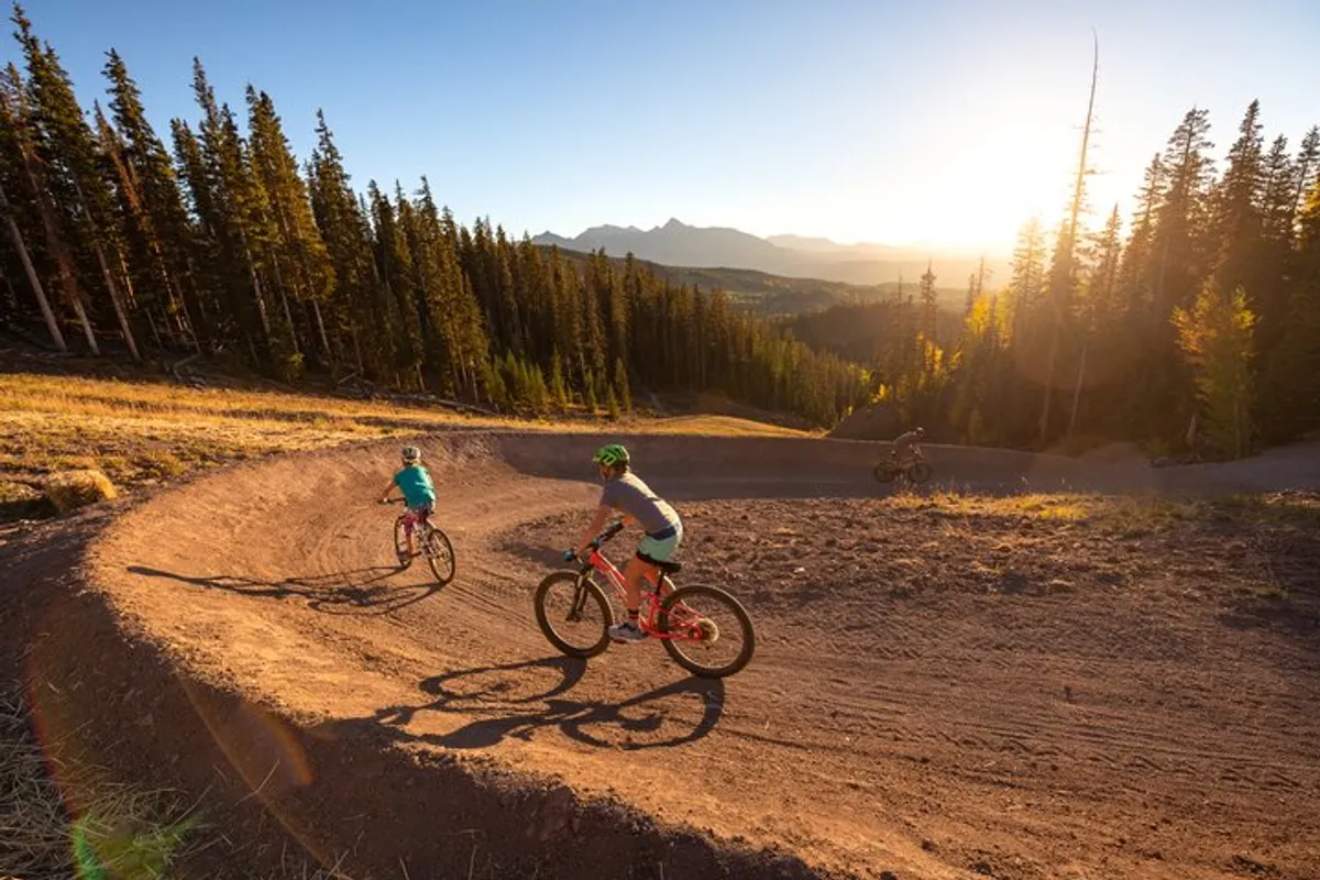 Mountain bikers carve along a winding trail flanked by tall pine trees, with a dramatic sunset skyline in the background. The scene encapsulates the thrill of outdoor adventure and the opportunity for friends or solo travelers to bond over exciting experiences in nature.