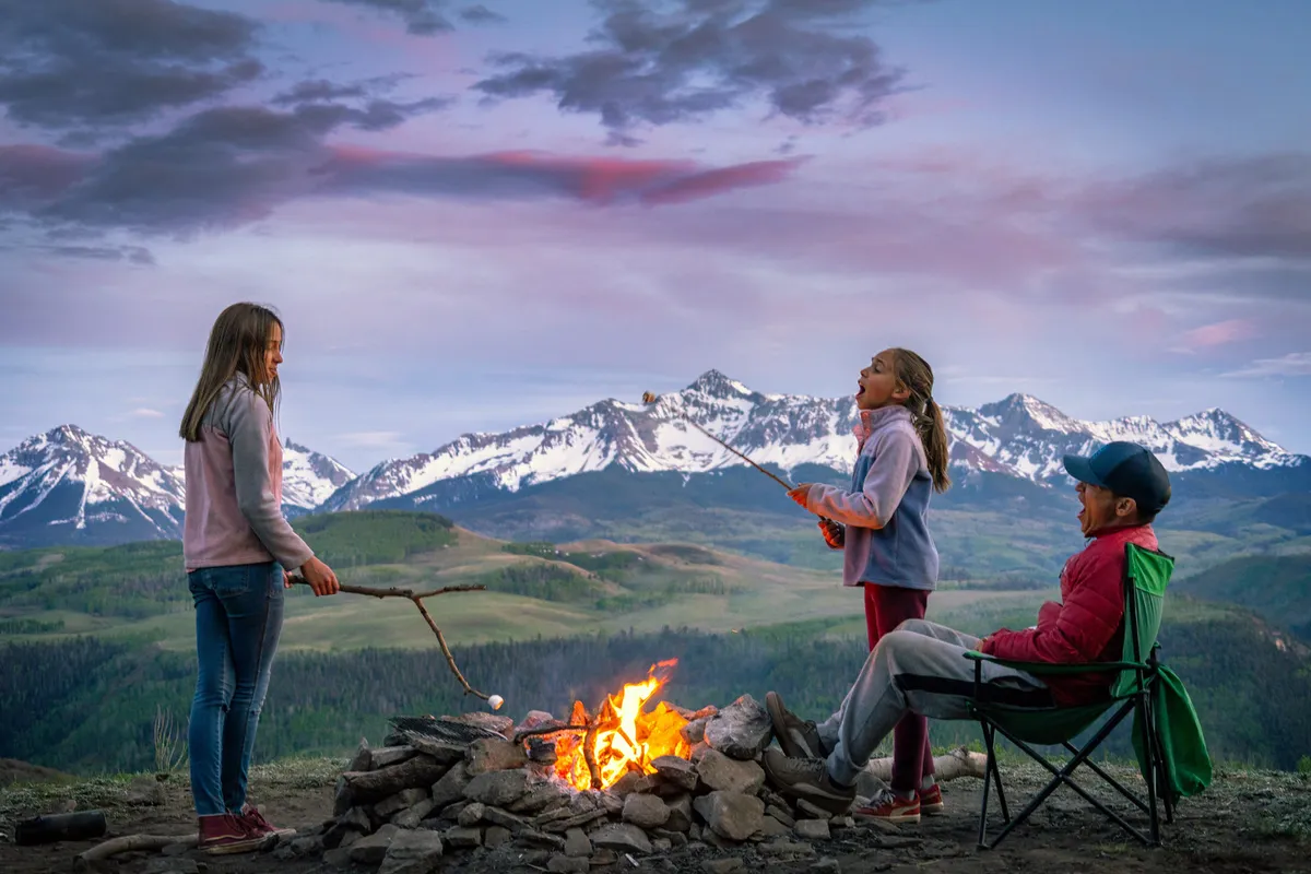 Children are gathered around a lively campfire, roasting marshmallows as dusk settles over a beautiful mountain range in the background. A relaxed adult sits in a chair nearby, enjoying the playful atmosphere and natural surroundings. Tall grasses and wildflowers add to the outdoor, family-friendly vibe.