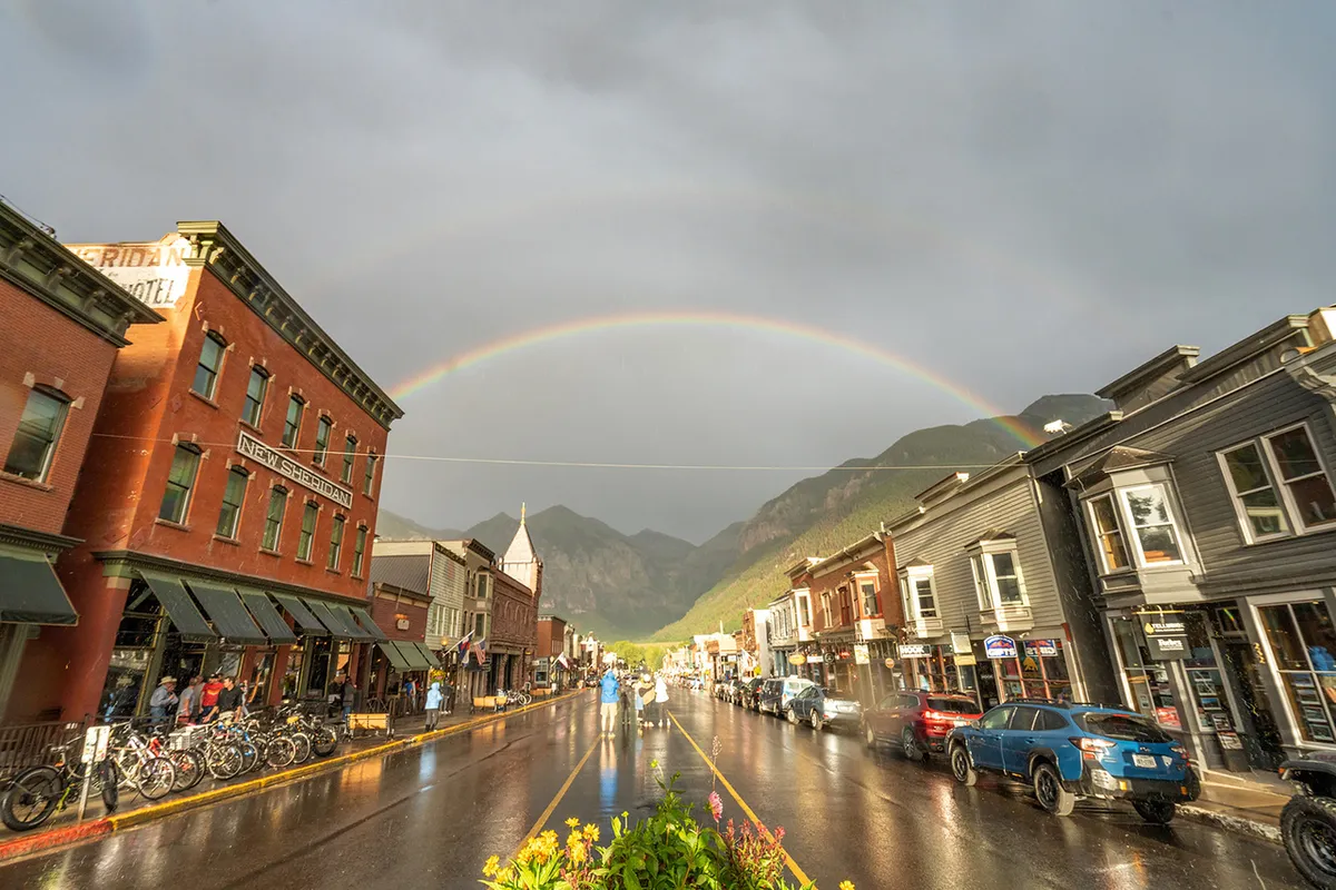 A vibrant view of Telluride’s historic downtown, with colorful buildings, a rainbow arching over wet streets, and bicycles parked along the sidewalk. The dramatic mountain backdrop sets the scene for walking tours, shopping, dining, and soaking up the town's unique charm.