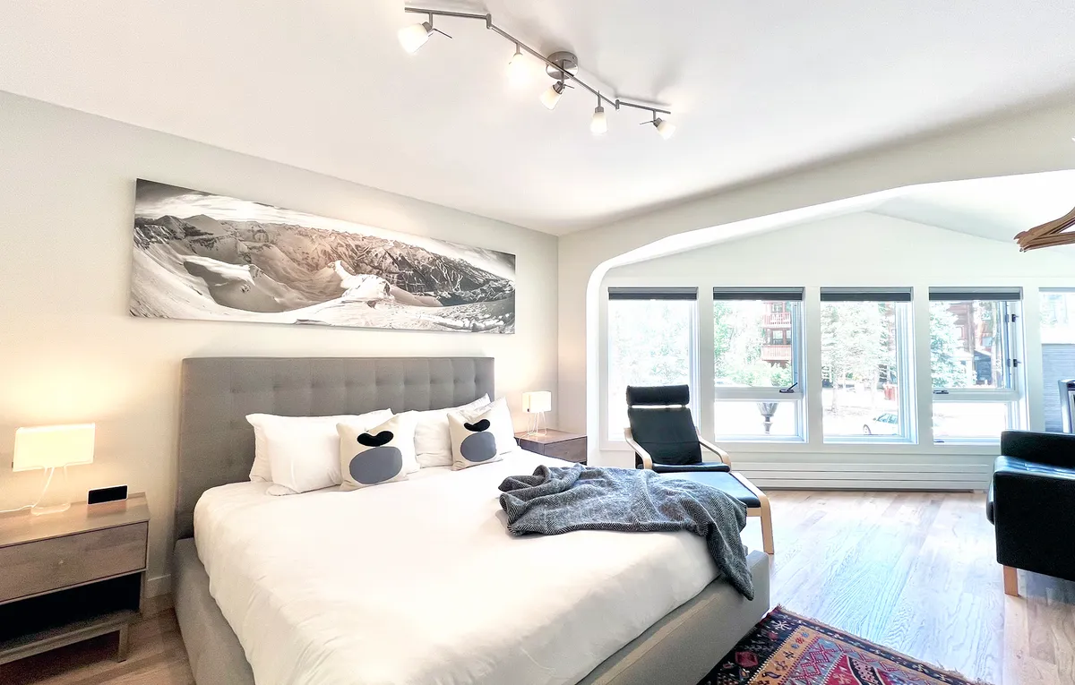 The image showcases a stylish, sunlit bedroom with a plush bed, modern mountain-themed artwork above the headboard, large windows letting in natural light, and a cozy armchair perfectly positioned for enjoying the stunning view. The tranquil space invites relaxation, with the Telluride mountain scenery visible just outside.