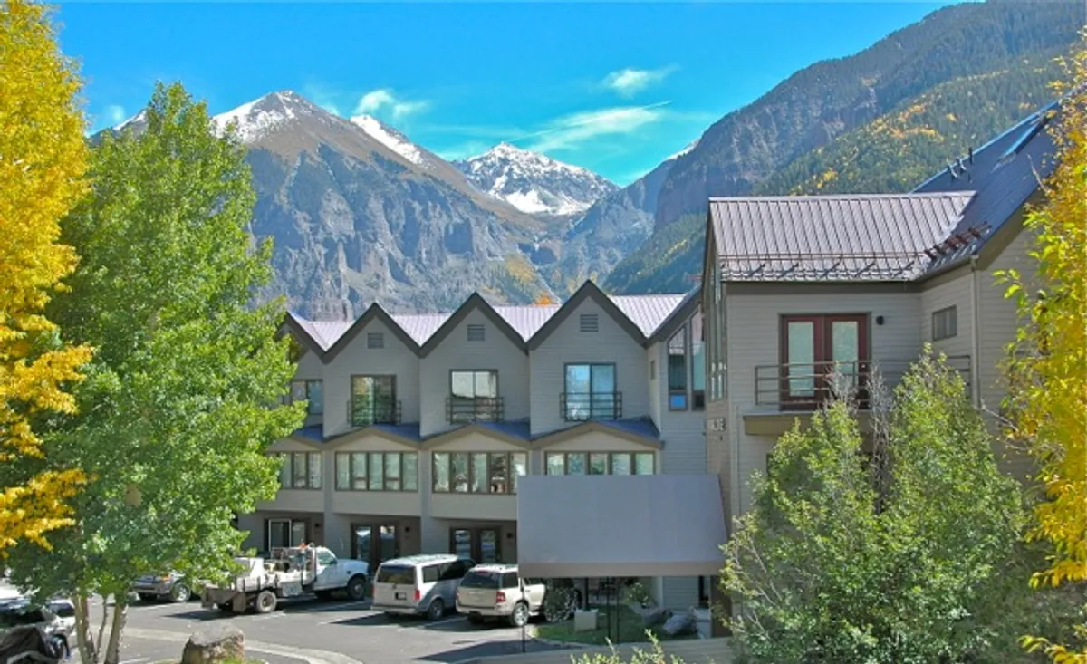 This image presents a spacious parking area in front of a modern building, set against a stunning mountain backdrop and highlighted by colorful autumn trees. The scene feels inviting and emphasizes ease of access, with ample outdoor space for gatherings or family activities.