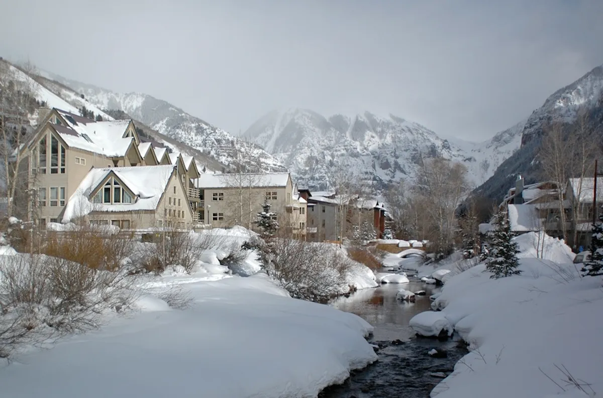 A stunning winter scene with snow-covered buildings beside a flowing river and majestic mountains in the background. The tranquil setting suggests cozy moments by the water after skiing or hiking, offering a quintessential Telluride atmosphere.