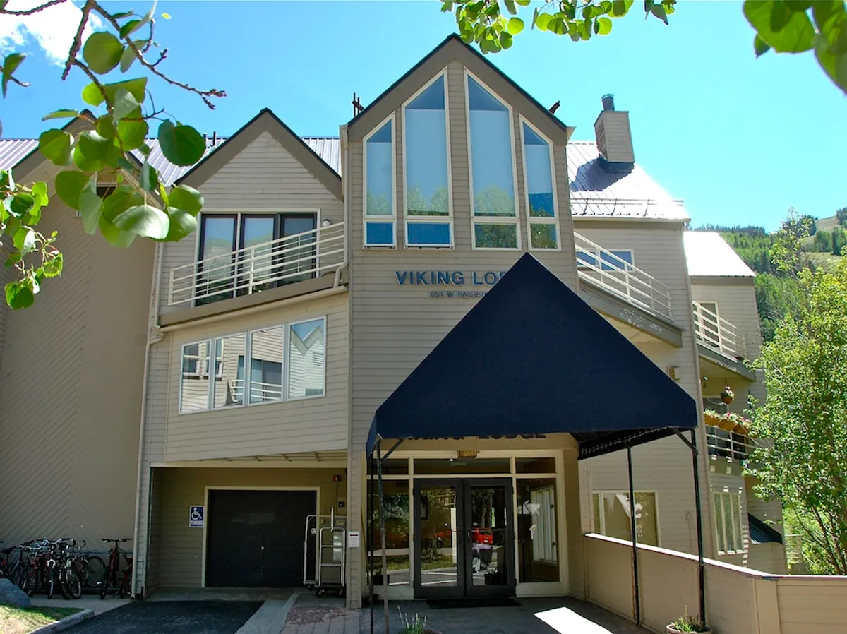 The image features the building’s large, angled windows and welcoming entrance awning. Balconies are visible, and people are seen socializing and biking, reinforcing a community-oriented, active lifestyle.