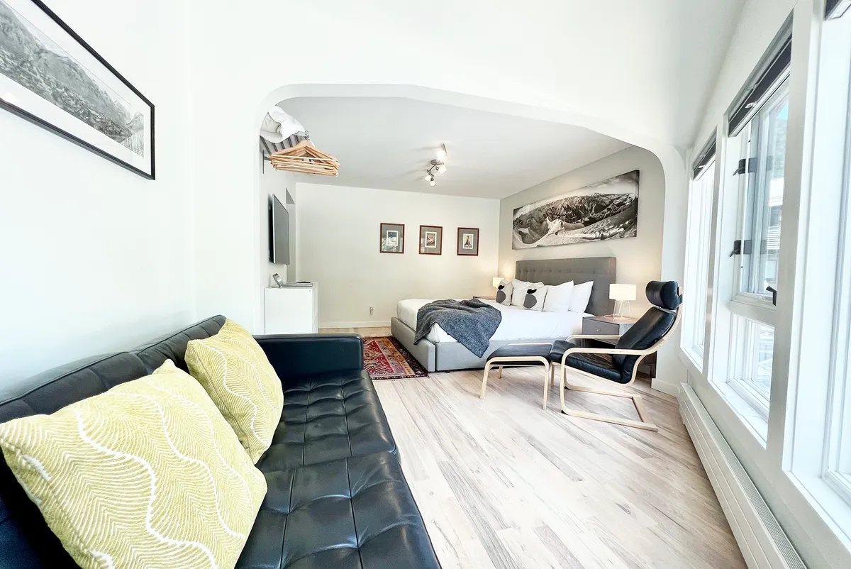 A beautifully lit living area with a stylish sofa adorned with decorative cushions, a bed with layered bedding, contemporary artwork, and large windows flooding the room with natural light. The scene embodies comfort and Scandinavian tranquility.
