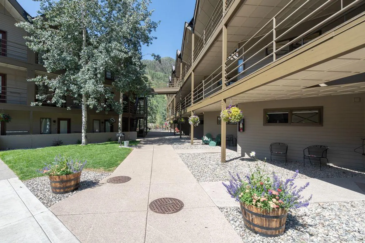 The exterior of a welcoming residential building is framed by lush gardens, flower planters, and mature trees, with balconies overlooking a walking path. People are seen strolling, relaxing on balconies, and enjoying the well-kept communal garden spaces, evoking a sense of comfort and togetherness.