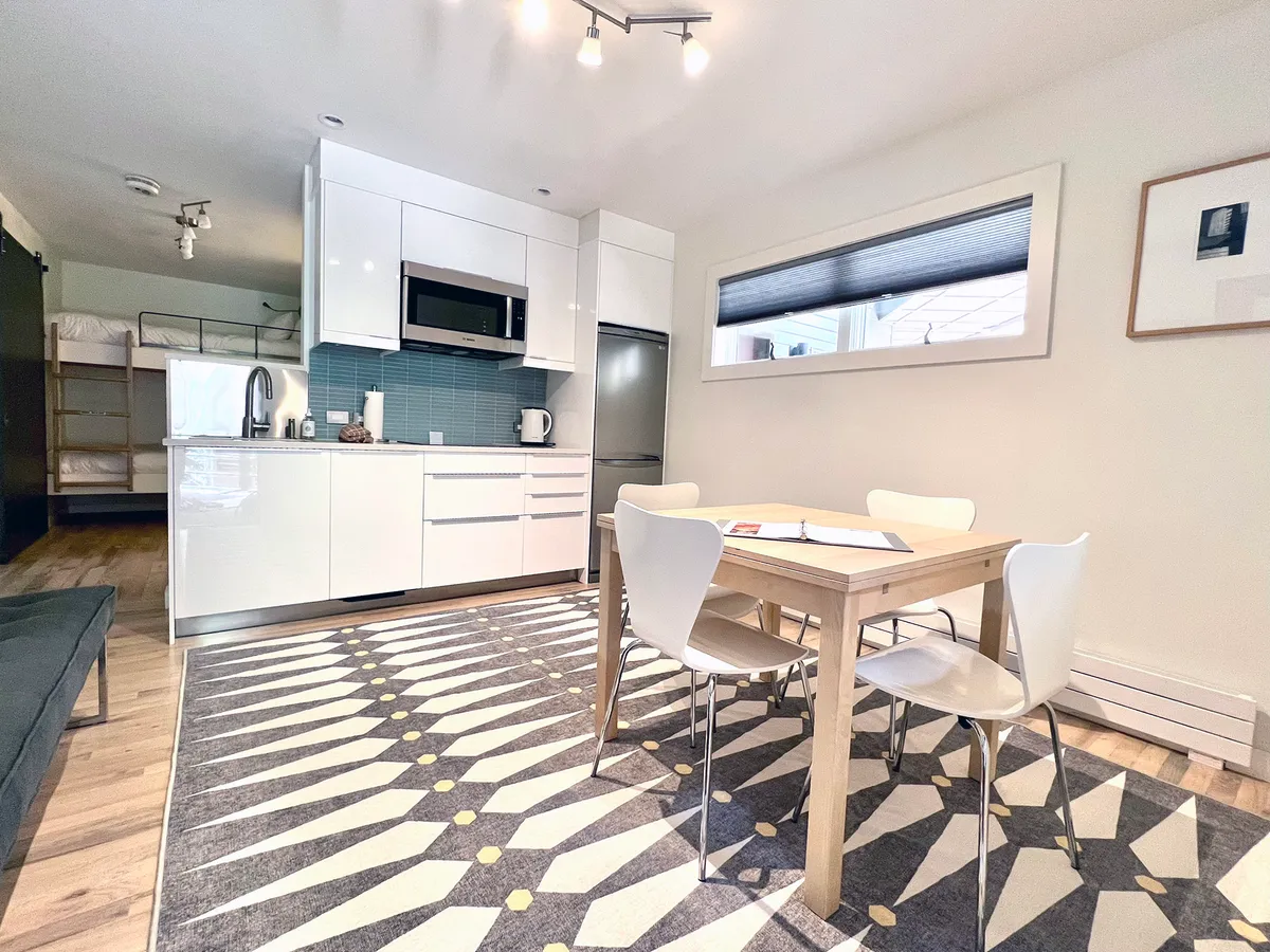 A modern, open-plan living and dining space featuring a dining table set for four, a compact kitchenette with sleek cabinetry and appliances, and a cozy rug underfoot. The area is bathed in natural light, giving a bright and airy feel ideal for unwinding or gathering after outdoor adventures.